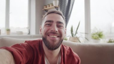 Göğüs hizasında, sakallı, koltukta oturan, akıllı telefonu uzanmış elinde tutan ve video görüşmesinde biriyle dostça sohbet eden beyaz bir adamın el kamerası görüntüsü.