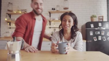 Mutfak masasında kahve fincanı, demlik ve video sohbetleriyle gülümseyen melez bir kadının göğüs fotoğrafı. Kocası geliyor, kameraya bakıyor ve neşeli bir şekilde konuşuyor.