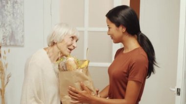 Genç Asyalı kadın bakıcı, neşeli yaşlı kadına bir torba yiyecek veriyor ve sonra onunla birlikte evde kameraya poz veriyor.