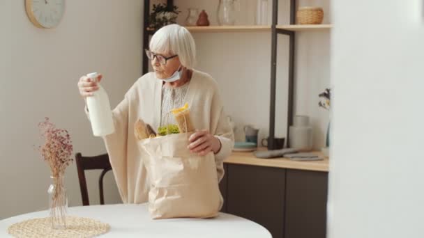 Senior Kvinna Med Skyddsmask Hakan Lossa Matvaror Från Papperspåse Köksbord — Stockvideo