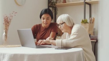 Genç Asyalı kadın bakıcı yaşlı beyaz bir kadınla masada oturuyor ve ona laptopu evde nasıl kullanacağını öğretiyor.