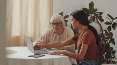 Evde dizüstü bilgisayar kullanmayı öğrenirken masada oturan ve genç bayan bakıcı ile konuşan yaşlı, gri saçlı kadının yan görüntüsü.