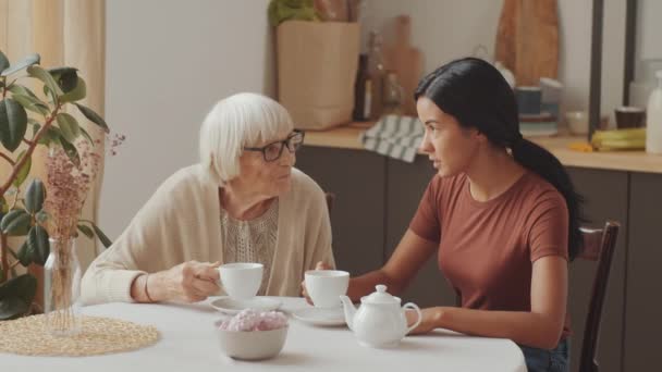 年轻的亚洲女性护理人员喝茶 在厨房桌上与年长的白人妇女交谈 — 图库视频影像