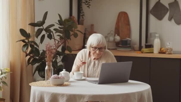 上了年纪的白种人女人 戴着眼镜坐在厨房桌旁 一边在笔记本电脑上打字 一边喝茶 一边在家里上网 — 图库视频影像