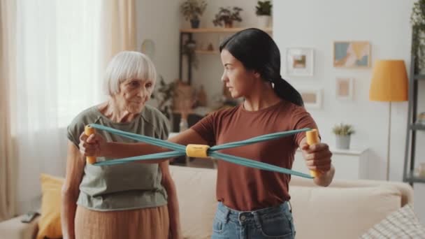 在亚洲女性护理人员的帮助下 年长的高加索妇女在家里接受训练的同时 与抵抗带一起进行武器锻炼 — 图库视频影像