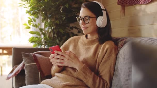 休日の家のソファに座っている間 スマートフォンを聞いて ワイヤレスヘッドフォンで若い陽気な女性のショットを傾けます — ストック動画
