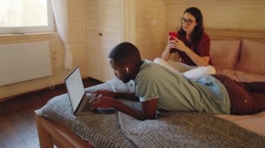 Kablosuz kulaklıklı genç Afro-Amerikalı adam evde yatıp laptopla çalışırken beyaz karısı akıllı telefonuyla mesajlaşıyor.