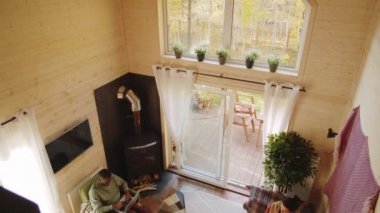 Karısı kanepede oturup kır evindeki akıllı telefondan internete bakarken Afro-Amerikan bir adamın laptopta çalışırken yüksek açılı görüntüsü.