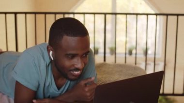 Kablosuz kulaklıklı genç Afro-Amerikalı adam evde yatağında yüzüstü yatıyor, gülümsüyor, el sallıyor ve dizüstü bilgisayardaki video konuşmasında konuşuyor.