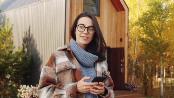 秋の日に森の中のコテージの前に屋外に立つ若い美しい女性とスマートフォンでメッセージ — ストック動画