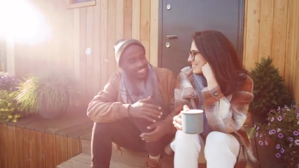 在阳光明媚的秋日 一位年轻快乐的高加索女人一边举着茶杯 一边微笑着与非洲裔美国丈夫聊天 一边坐在度假屋的门廊上 拍下了一张弧形的照片 — 图库视频影像