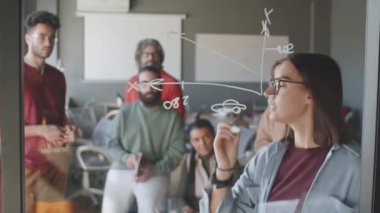 Genç ve güzel bir kadın cam duvara işaretleyici ile grafik çiziyor ve ofis toplantısında çok ırklı iş arkadaşlarından oluşan bir ekibe iş planını anlatıyor.
