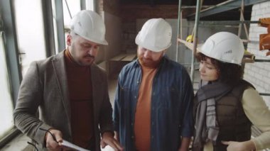 Miğferli bir erkek mimarın yarım kalmış bir dairede dikilip danışmanlık yaparken beyaz aile çiftine panodaki bir şeyi açıklarken çekilen görüntüsü.