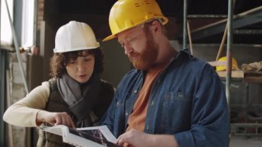 İnşaat alanında iş birliği yaparken erkek meslektaşımla inşaat planlarını tartışan kasklı genç bir kadın mimarın göğüs yukarı çekimi.