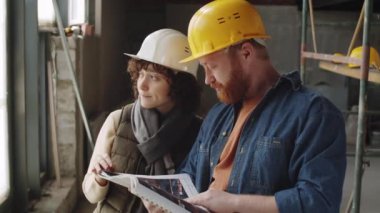 Erkek ve kadın mimarların kasklarıyla göğüs göğüse çekilip tartıştıktan sonra birlikte kameraya poz verirken ve daralma alanında çalışırken gülümserken.