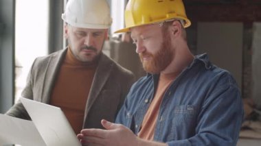 İnşaat alanında birlikte çalışırken laptopta bir şey tartışan kasklı iki erkek mimarın göğüs görüntüsü.