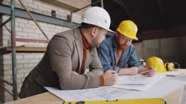 Kafkas şapkalı iki beyaz erkek mühendis çalışma masasına yaslanıyor ve inşaat alanında birlikte çalışırken kat planı hakkında konuşuyorlar.