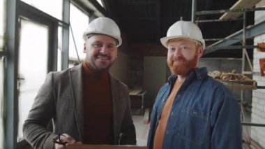 Miğferli mimar ve erkek müşterinin belden yukarı portresi. Yeni tamamlanmamış apartman dairesinde dikilip kameraya bakıyor ve gülümsüyor.