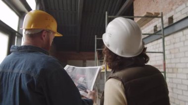 Erkek ve kadın mühendislerin yarım kalmış apartman dairesinde dikilip inşaat projesinin kağıt slaytlarını tartışmalarının arka plan görüntüsü