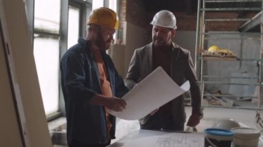 Kask takmış iki erkek mimar, inşaat alanında çalışırken kat planlarını inceliyor ve birlikte konuşuyorlar.