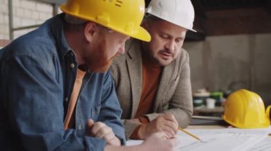 İki erkek mimar masaya yaslanıyor ve tamamlanmamış bir binada birlikte çalışırken kağıt üzerinde inşaat planını tartışıyorlar.