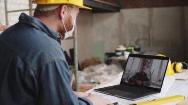 Koruyucu yüz maskesi takan erkek mimar inşaat alanında erkek meslektaşıyla uzaktan çalışırken bilgisayardan video bağlantısı kuruyor.