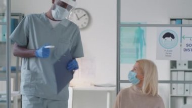 Koruyucu maske, ekran ve eldiven takan Afro-Amerikalı tıp çalışanı. Kvid-19 salgını sırasında doktorla görüşmeden önce kızıl ötesi termometresi olan kadın hastanın vücut ısısını kontrol ediyor.