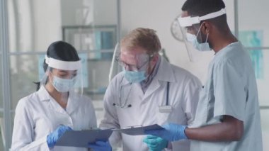 Tıbbi üniformalı, koruyucu maskeli, ekranlı ve eldivenli çok ırklı erkek ve kadın doktorlar klinikte covid-19 salgını sırasında birlikte çalışırken panoda dokümanları tartışıyorlar.
