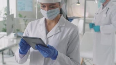 Laboratuvar önlüğü, yüz ekranı ve maske takmış genç Asyalı kadın doktorların podyum 19 salgını sırasında dijital tablet kullanırken görüntülerini yukarı kaldır.