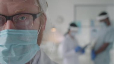 Korunaklı yüz kalkanı ve maskesiyle Coronavirus salgını sırasında tıbbi ofiste çalışırken kameraya bakan kendine güvenen erkek doktoru yakından çek.