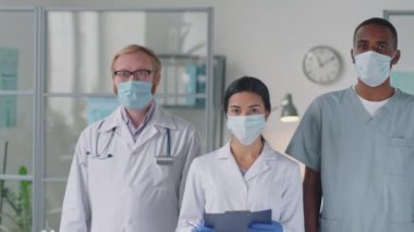 Profesyonel üniformalı ve koruyucu maskeli çeşitli doktorların portresi tıbbi ofiste yan yana duruyor ve Coronavirus salgını sırasında kameraya poz veriyor.