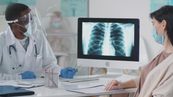 Afro-American doctor in lab coat, protective face screen, mask and gloves showing lunge x-ray on computer and talking to female patient in medical office during covid-19 pandemic — Stock Footage Afro American Doctor Lab Coat Protective Face Screen Mask Gloves Stock Video