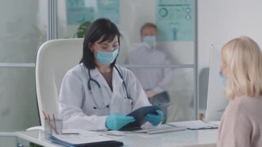 Laboratuvar önlüklü, maskeli ve eldivenli profesyonel kadın doktor tıbbi geçmişi kontrol ediyor ve klinikte muayene sırasında kadınla konuşuyor.