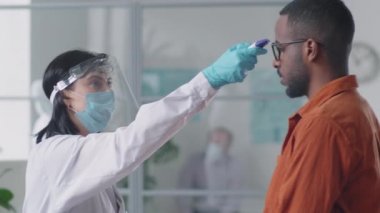 Koruyucu maskeli, ekranlı ve eldivenli kadın doktor kızılötesi termometresi olan siyahi erkek hastanın sıcaklığını ölçerken tıbbi muayenede kovid-19 kontrolü yapıyor.