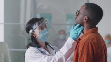 Laboratuvar önlüklü kadın doktor, koruyucu yüz maskesi, ekran ve eldivenler klinikte Afro-Amerikalı bir adamın boynunu ve boğazını incelerken Coperid-19 kontrolü yapıyor.