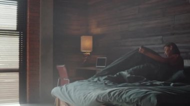 Genç Asyalı bir kadının yataktan kalktığını, panjurlarla pencereye doğru yürüdüğünü ve sabahları akıllı telefondan yazdığını gösteren bir fotoğraf.