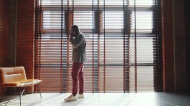 Genç Afro-Amerikalı iş adamının tavan arasında geniş bir pencerenin yanında yürürken ve cep telefonuyla konuşurken tam boy görüntüsü.