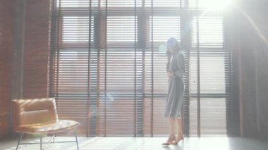Genç ve şık bir iş kadınının pencerenin yanında perdelerle yürürken ve çatı katında cep telefonuyla konuşurken görüntüsünü yakınlaştır.