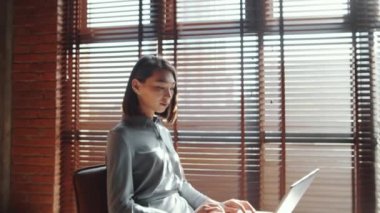 Çekici Asyalı genç bir kadının portre fotoğrafını yakınlaştır. Dizüstü bilgisayar kullanarak ve tavan arasında güneş ışığıyla dolu bir odada kameraya bakarken.