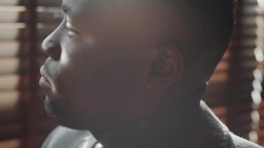 Kendine güvenen Afro-Amerikalı iş adamının pencerenin yanında panjurlarla otururken kameraya baktığı el kamerası görüntülerini kapatın.