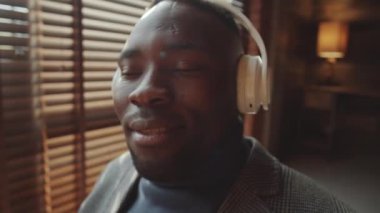 Kablosuz kulaklıklı, müzik dinleyen ve merkez ofisindeki kameraya poz veren genç Afro-Amerikan bir adamın yakın çekim görüntüsü.