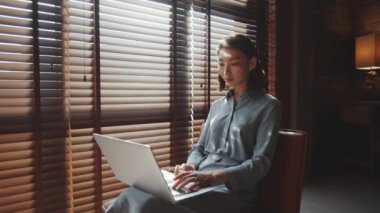 Genç ve çekici Asyalı iş kadınının, ofis panjurlarıyla pencerenin yanında otururken ve dizüstü bilgisayarla çalışırken görüntüsünü büyüt.