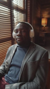 Kablosuz kulaklıklı genç bir adamın, zevkle müzik dinlerken, ritmi ayarlarken ve ofiste otururken kameraya bakarken dikey görüntüsü.