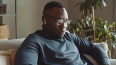 Genç siyahi adamın kablosuz kulaklıklarıyla kanepede otururken ve dizüstü bilgisayarda yazarken yakın plan fotoğrafını çek.