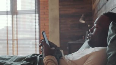 Genç Afro-Amerikalı bir adamın sabah evde yatakta yatarken akıllı telefondan mesajlaşmasını yakından çek.