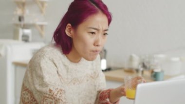 Pembe saçlı, portakal suyu içen ve evde kahvaltı yaparken dizüstü bilgisayarda yazan genç Asyalı kadın.