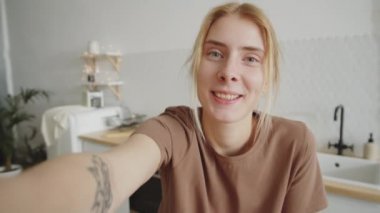 Genç, neşeli, sarışın bir kadın evde oturuyor. Uzamış koluyla akıllı telefonu tutuyor. Kameraya bakıyor ve online video görüşmesinde sohbet ediyor.