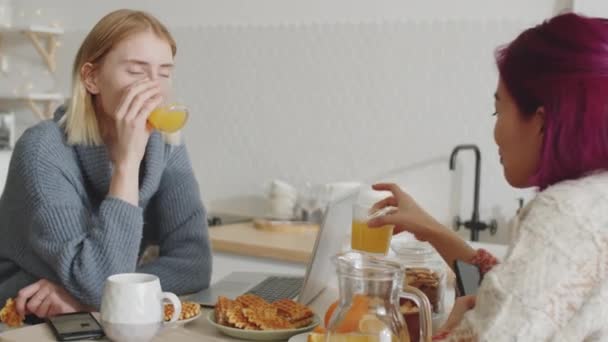 年轻的白人和亚裔女性室友们坐在厨房桌旁 喝着橙汁 吃着华夫饼 早上在家里聊天 — 图库视频影像