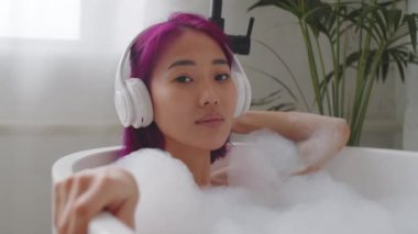 Küvette uzanmış pembe saçlı çekici bir Asyalı kadın kablosuz kulaklıkla müzik dinliyor, kameraya bakıyor ve gülümsüyor.