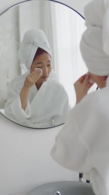 Bornoz ve havlu giymiş genç Asyalı bir kadının kafasına pamuk pedlerle losyon sürerken ve banyodaki aynaya bakarken dikey pozu.